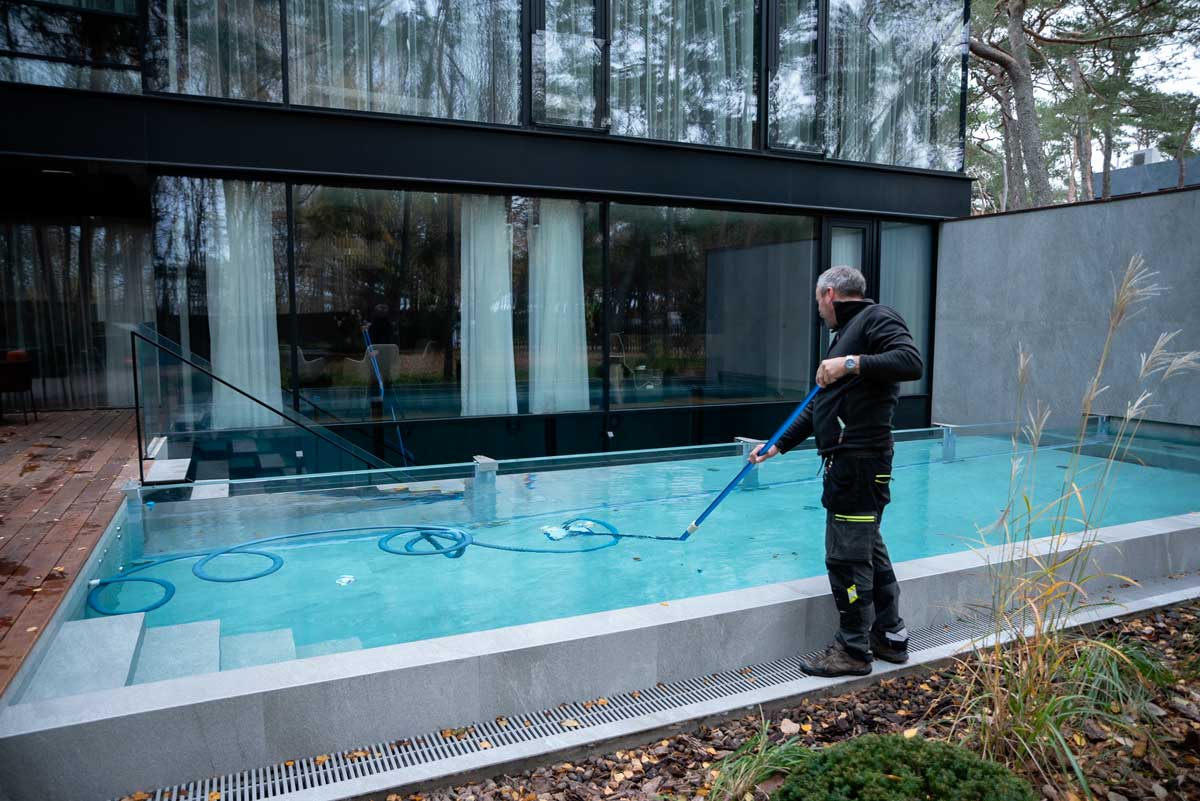 Piscina al aire libre con agua turbia y sucia, manchas marrones y suciedad visibles en la superficie, una persona de pie junto a la piscina.