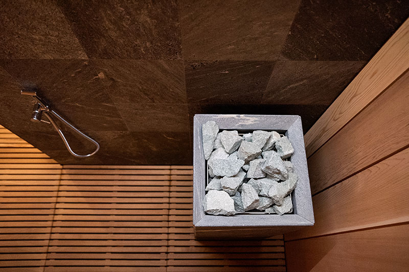 Vista superior de una estufa de sauna con piedras de color gris claro, rejillas de madera y azulejos de pared oscuros.