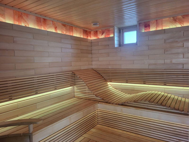 Sauna de madera clara con banco ondulado e iluminación LED.