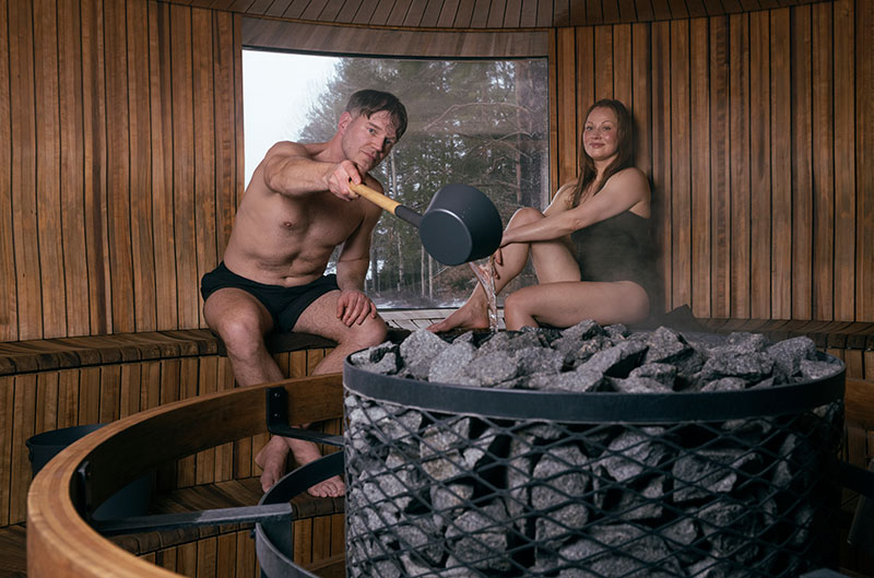 Un hombre vierte agua sobre las piedras de sauna calentadas con un cucharón, una mujer está sentada cerca; una sauna de madera humeante, redonda y con ventana.
