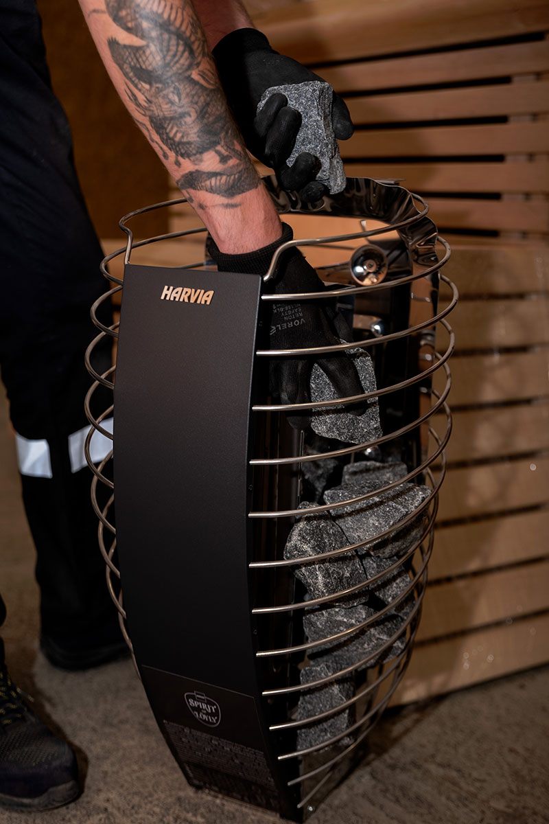 Primer plano de la cesta de una estufa eléctrica HARVIA: piedras de sauna de color gris oscuro se colocan en guantes negros.