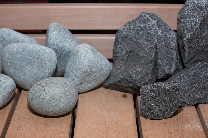 Comparación: piedras de río redondeadas de color claro y piedras de sauna rotas y oscuras colocadas sobre un banco de madera.